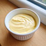 A small white ramekin filled with thick, pale yellow Vegan Creamy Lemon Tahini Dressing, showing swirls on the surface.