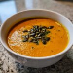 Close-up of a vibrant orange Vegan Creamy Roasted Carrot Ginger Soup topped with toasted pumpkin seeds.