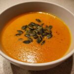 Close-up of a white bowl filled with bright orange Vegan Creamy Roasted Carrot Ginger Soup, topped with dark green pumpkin seeds.