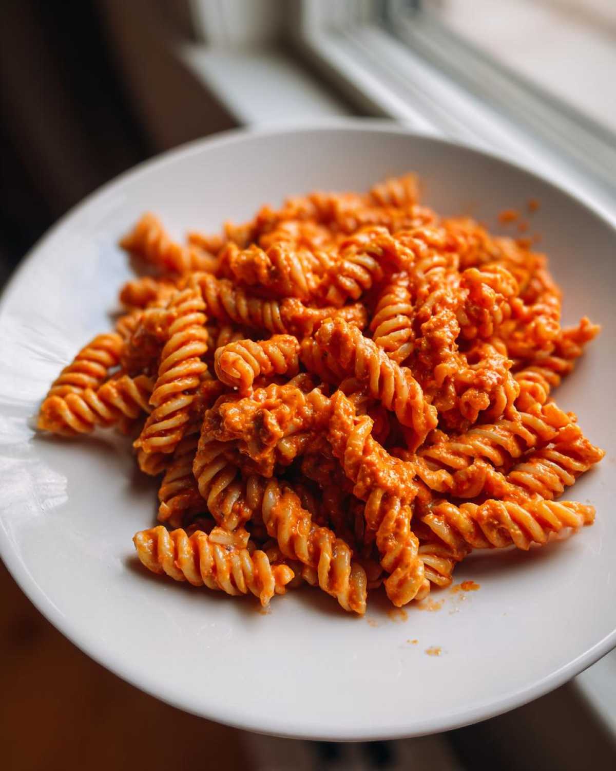 Close-up of fusilli pasta coated in a rich, orange Vegan Creamy Roasted Red Pepper Hummus Pasta sauce, served on a white plate.