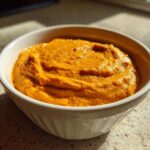 A white bowl filled with vibrant orange Vegan Creamy Sweet Potato Mac Sauce, sitting on a speckled countertop.