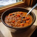 A close-up of a dark bowl filled with thick Vegan One-Pot Lentil Taco Soup, featuring lentils, black beans, and chunks of carrot.
