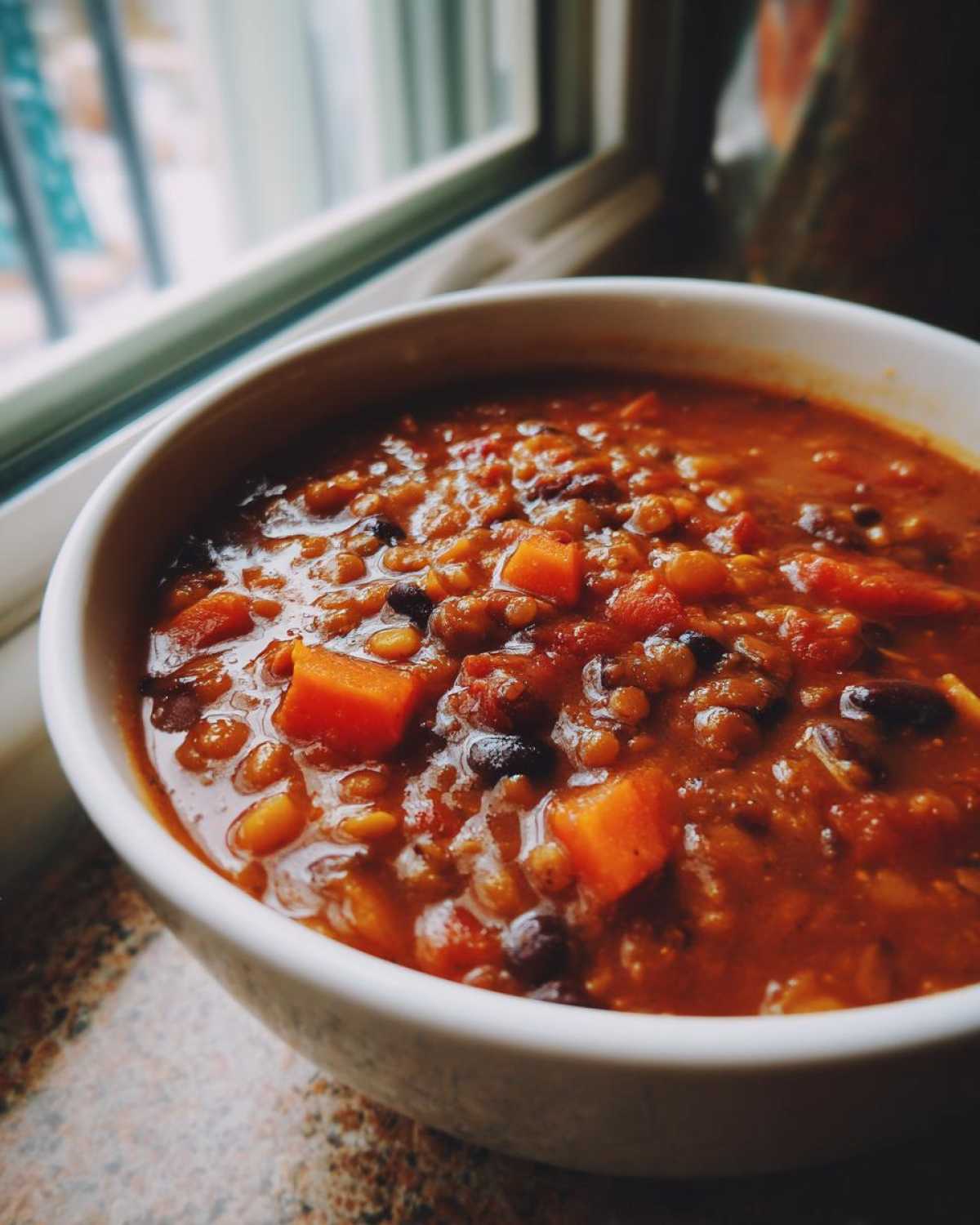 Close-up of a bowl filled with rich, red Vegan One-Pot Lentil Taco Soup featuring lentils, black beans, and diced orange vegetables.