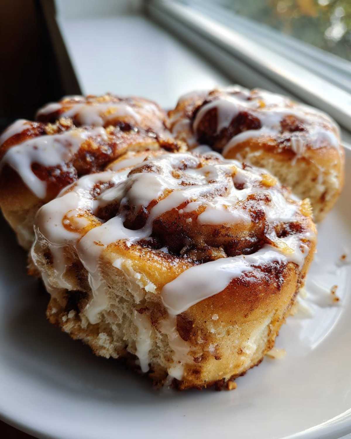Close-up of three gooey Banana Bread Cinnamon Rolls Dessert drizzled with white icing on a white plate.