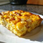 A golden-brown slice of Cheesy Egg Casserole topped with visible pieces of cooked onion, served on a white plate.