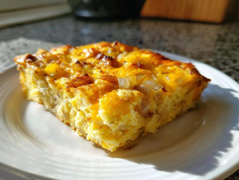 A golden-brown slice of Cheesy Egg Casserole topped with visible pieces of cooked onion, served on a white plate.