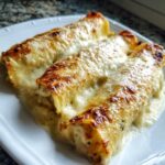 Three baked rolls of Chicken Manicotti Alfredo covered in bubbly, browned Alfredo sauce and melted cheese.