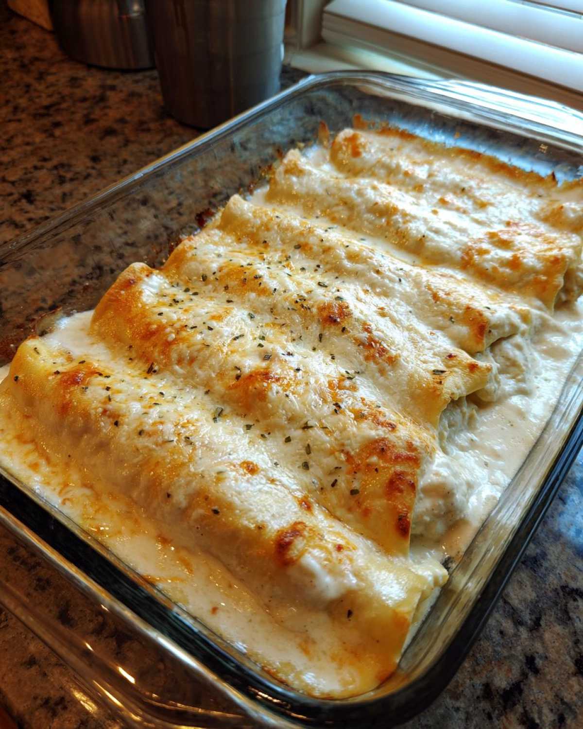 Close-up of baked Chicken Manicotti Alfredo covered in melted, browned cheese and Alfredo sauce in a clear baking dish.