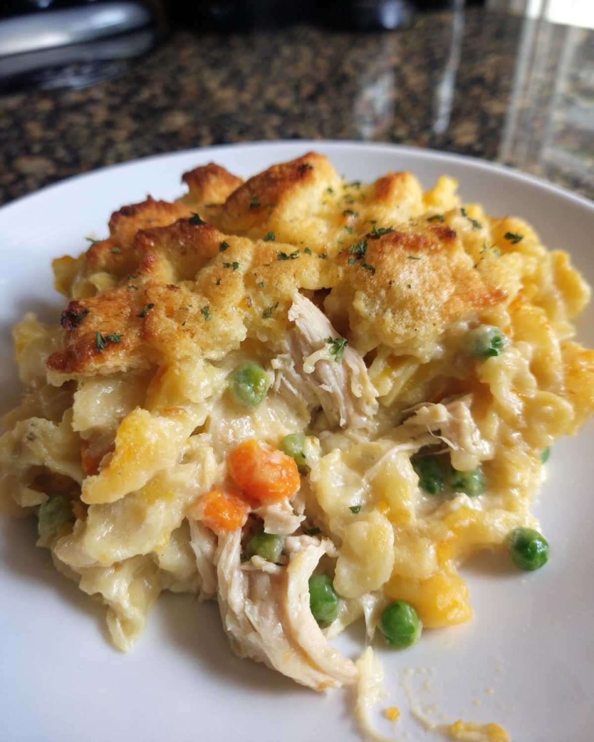 A close-up serving of creamy Chicken Noodle Casserole featuring shredded chicken, peas, carrots, and a golden, biscuit-like topping.