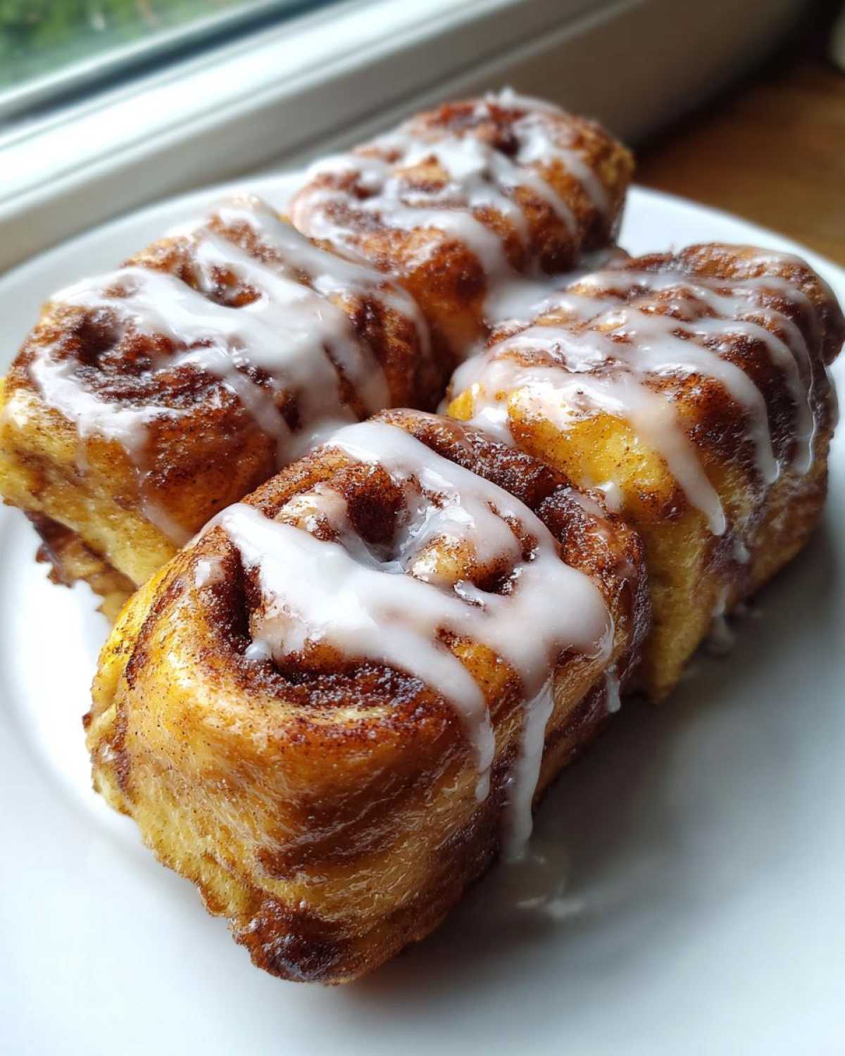 Four golden Cinnamon Roll French Toast Roll Ups drizzled generously with white vanilla icing on a white plate.