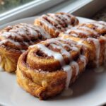Four golden Cinnamon Roll French Toast Roll Ups drizzled generously with white sweet icing on a white plate.
