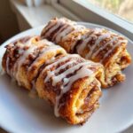 Four golden brown Cinnamon Roll French Toast Roll Ups drizzled generously with white icing on a white plate.