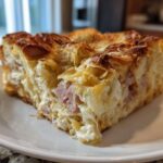 A close-up of a square slice of Cozy Croissant Breakfast Bake featuring flaky pastry, ham, and melted cheese on a white plate.