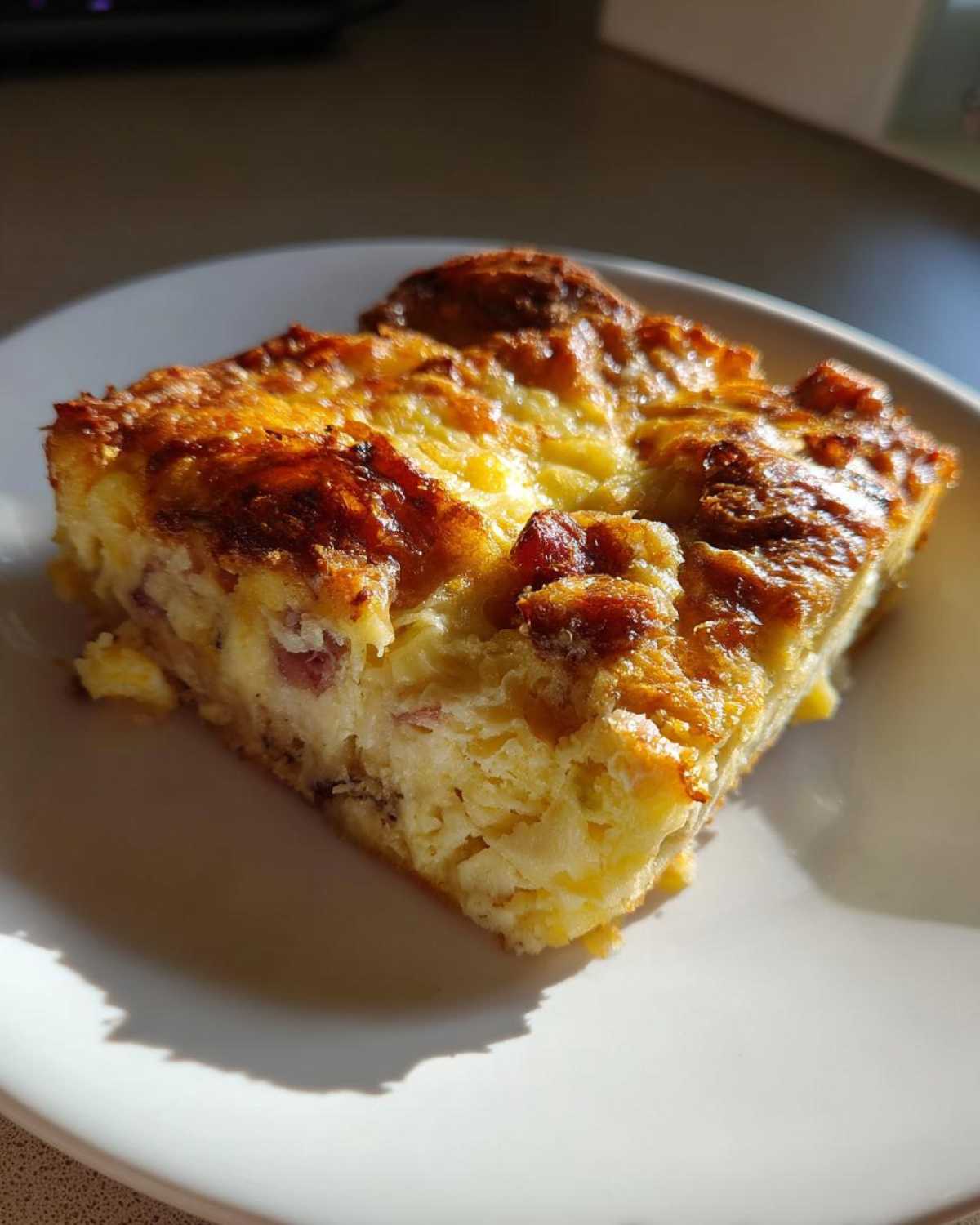 A square slice of Cozy Croissant Breakfast Bake with a golden-brown, cheesy top, served on a white plate.