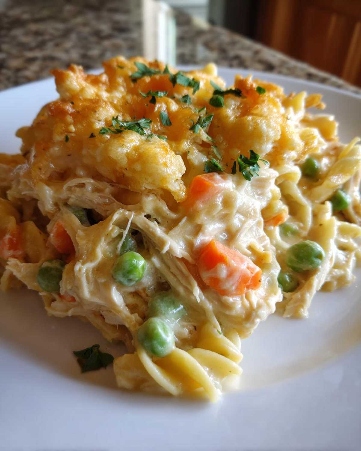 A close-up of a serving of creamy Chicken Noodle Casserole featuring shredded chicken, peas, carrots, and a golden, baked topping garnished with parsley.