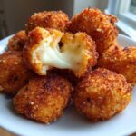 A pile of golden brown Crispy Fried Mozzarella Bites on a white plate, one is broken open showing melted, stretchy cheese.