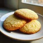Three golden brown Flavor Nectar Sugar Cookies stacked slightly on a light-colored plate near a window.