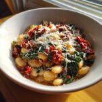 A close-up of a bowl of Marry Me White Beans topped with shredded Parmesan cheese, sun-dried tomatoes, and wilted greens.
