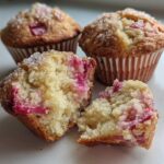 A close-up of Moist Sour Cream Rhubarb Muffins, one broken open showing the moist crumb and pink rhubarb pieces.