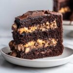 A rich slice of Reese Peanut Butter Cup Earthquake Cake Chocolate showing three dark chocolate layers filled with peanut butter frosting and candy pieces.