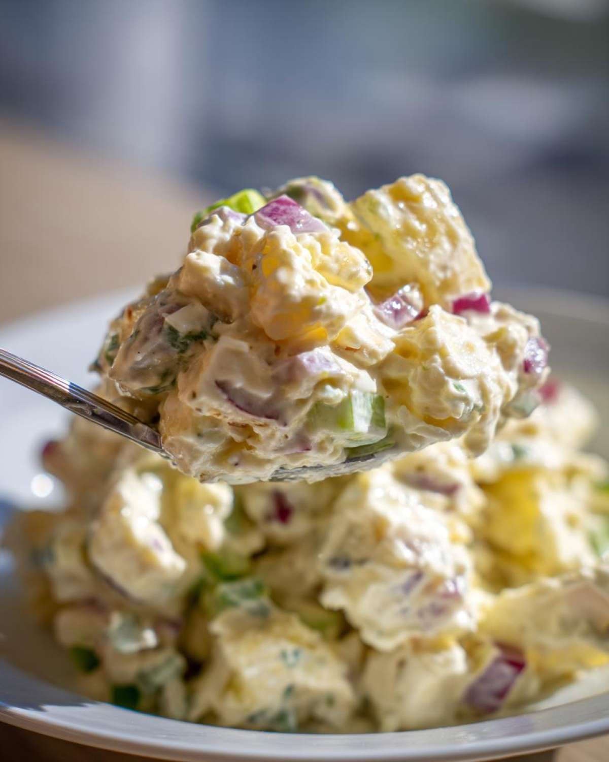 A spoonful of creamy potato salad, featuring chunks of potato, red onion, and celery, lifted from a white bowl.