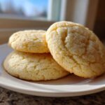 Three freshly baked Sugar Cookies - Flavor Nectar, coated in sugar, stacked on a small white plate near a sunny window.